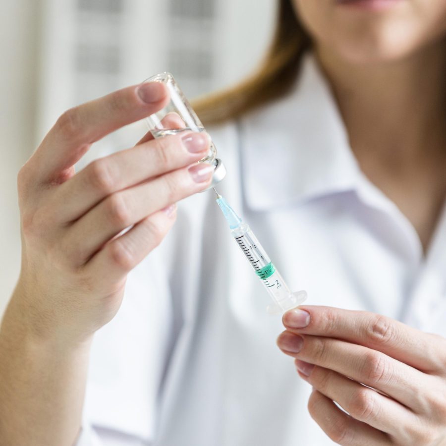 defocused-female-scientist-with-safety-glasses-holding-syringe-with-vaccine defocused-female-scientist-with-safety-glasses-holding-syringe-with-vaccine